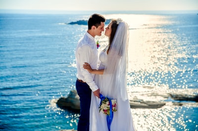 女人在结婚礼服吻男人在白色礼服衬衫拿束花在白天附近的水域
